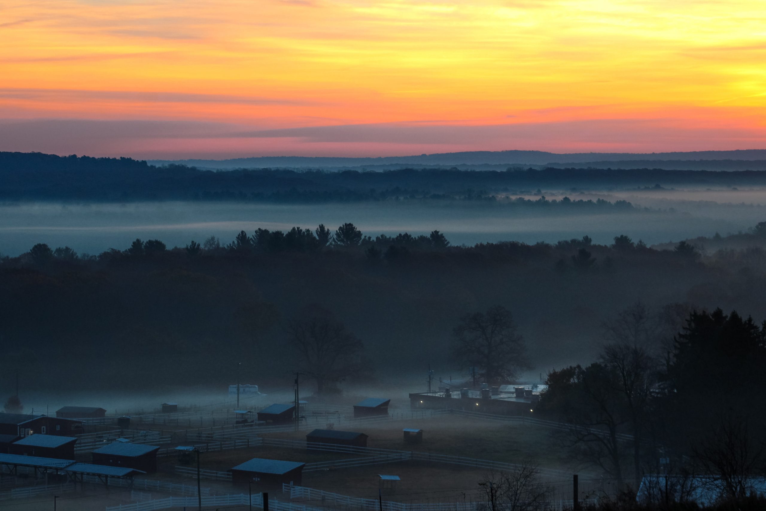 A foggy sunrise over rolling hills and farmland, with soft orange and blue light spreading across the sky.