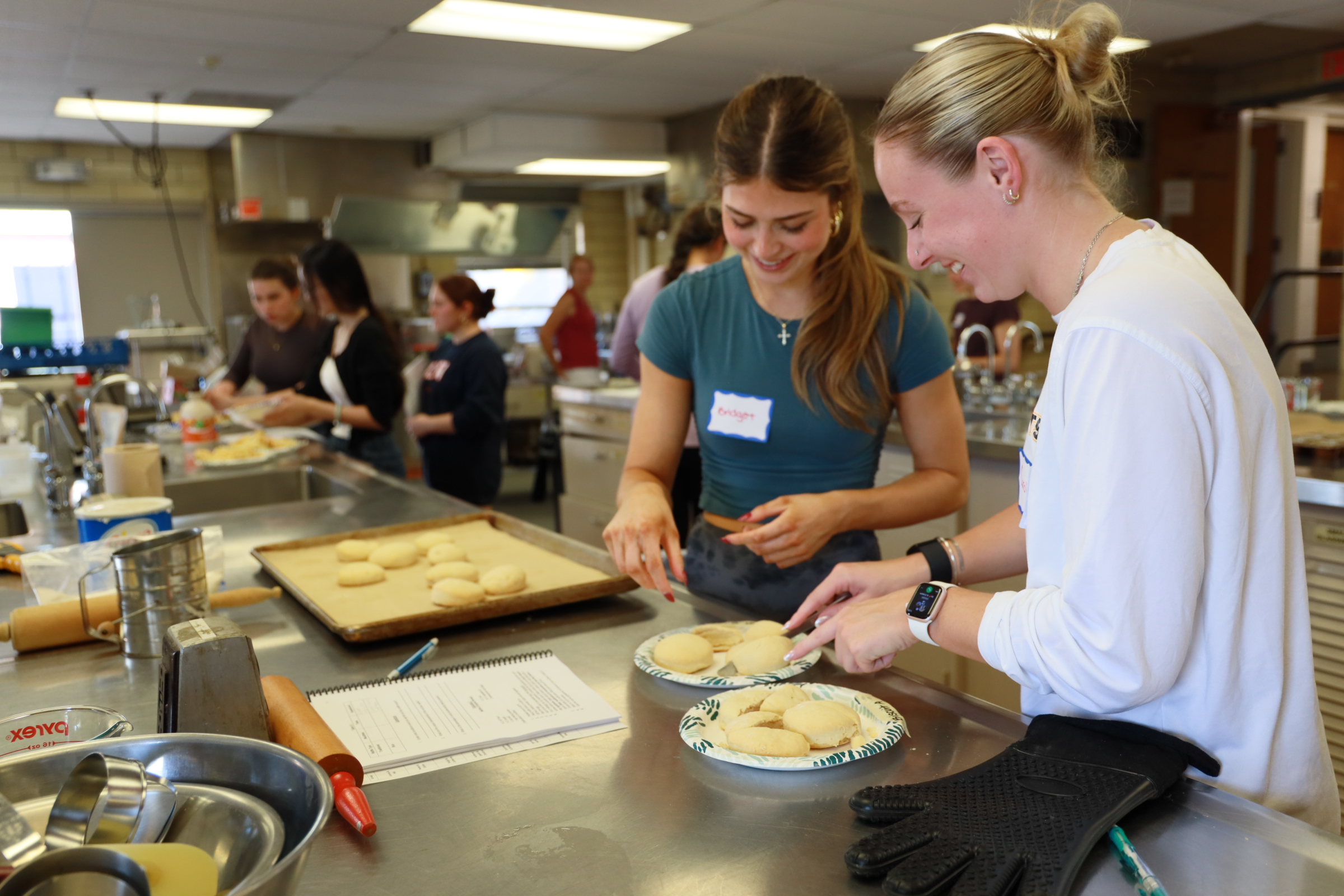 Students in a kitchen classroom smile while preparing and shaping dough on plates and trays during a bread-making activity.