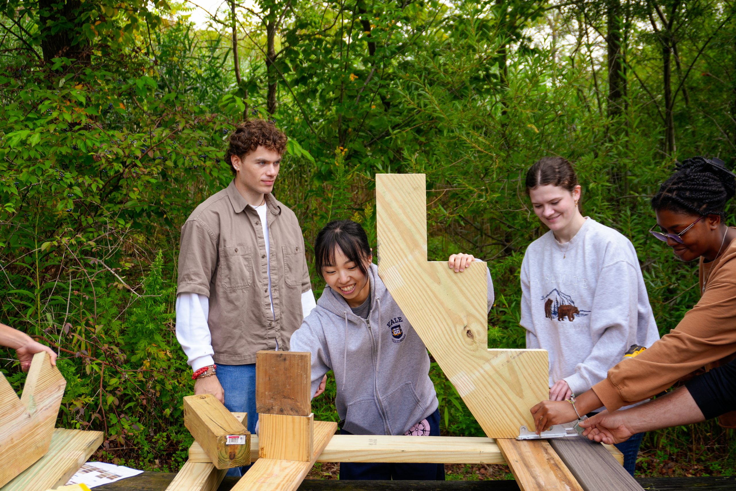 Four students work together outdoors to assemble a wooden structure, smiling as they measure and position the boards amid green foliage.