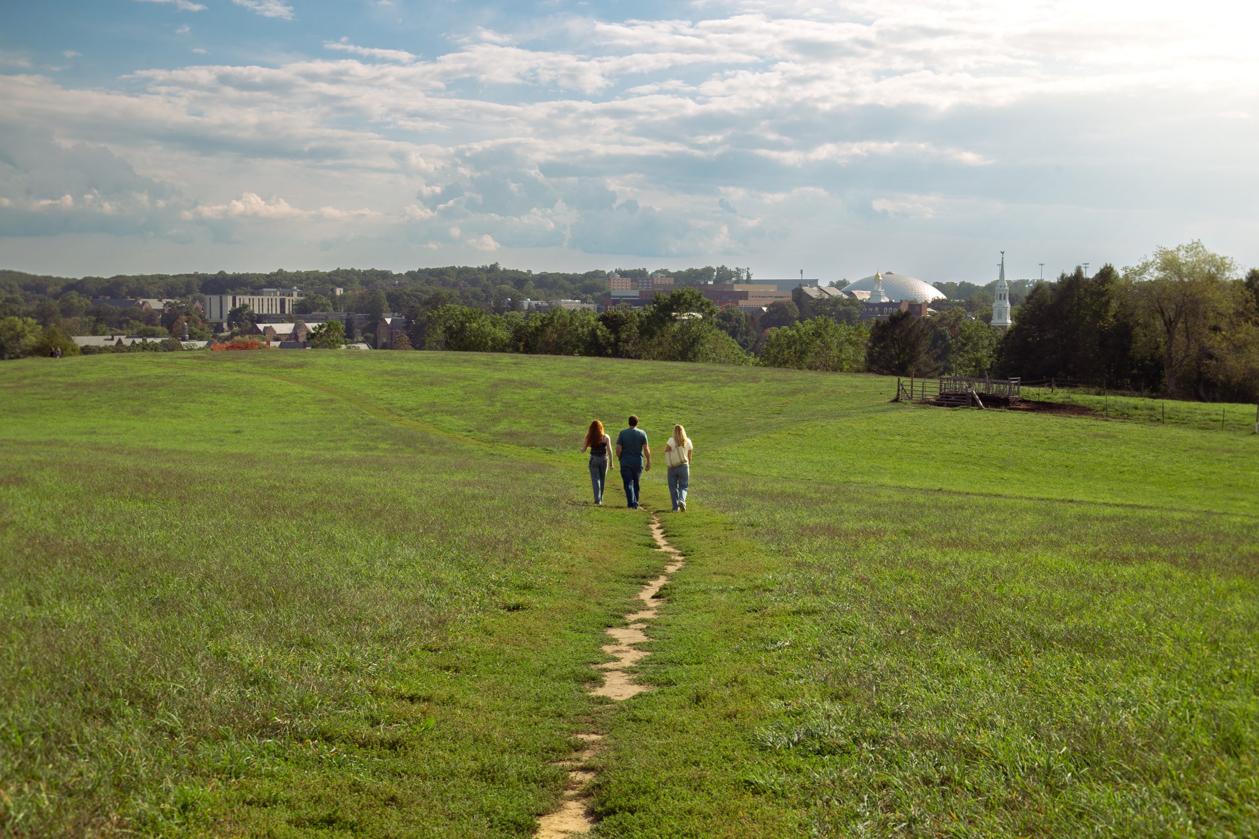 Three people walking along a narrow dirt path through a wide green field toward a distant campus under a partly cloudy sky.